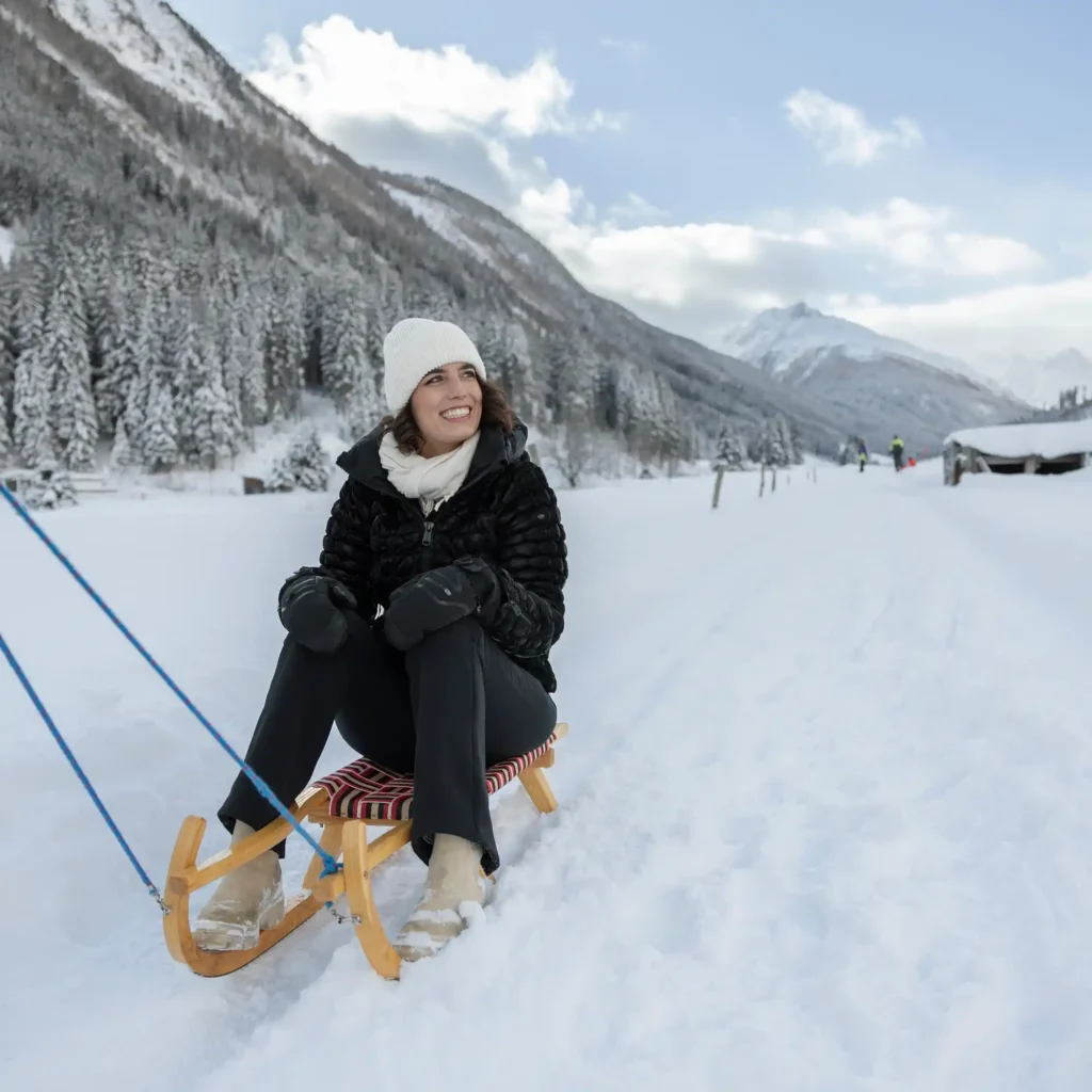 A woman in winter clothing sits on a wooden sled in a snowy landscape, smiling. She wears a white beanie, black jacket, gloves, and dark pants. Snow-covered trees and hills are in the background under a partly cloudy sky.