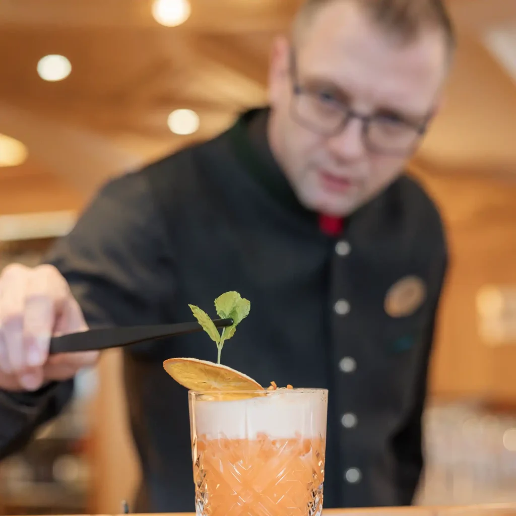 A bartender in a black uniform meticulously places a garnish on a frothy orange cocktail in a textured glass. The background is softly blurred, highlighting the focus on the drink and the bartenders precise hand gesture.