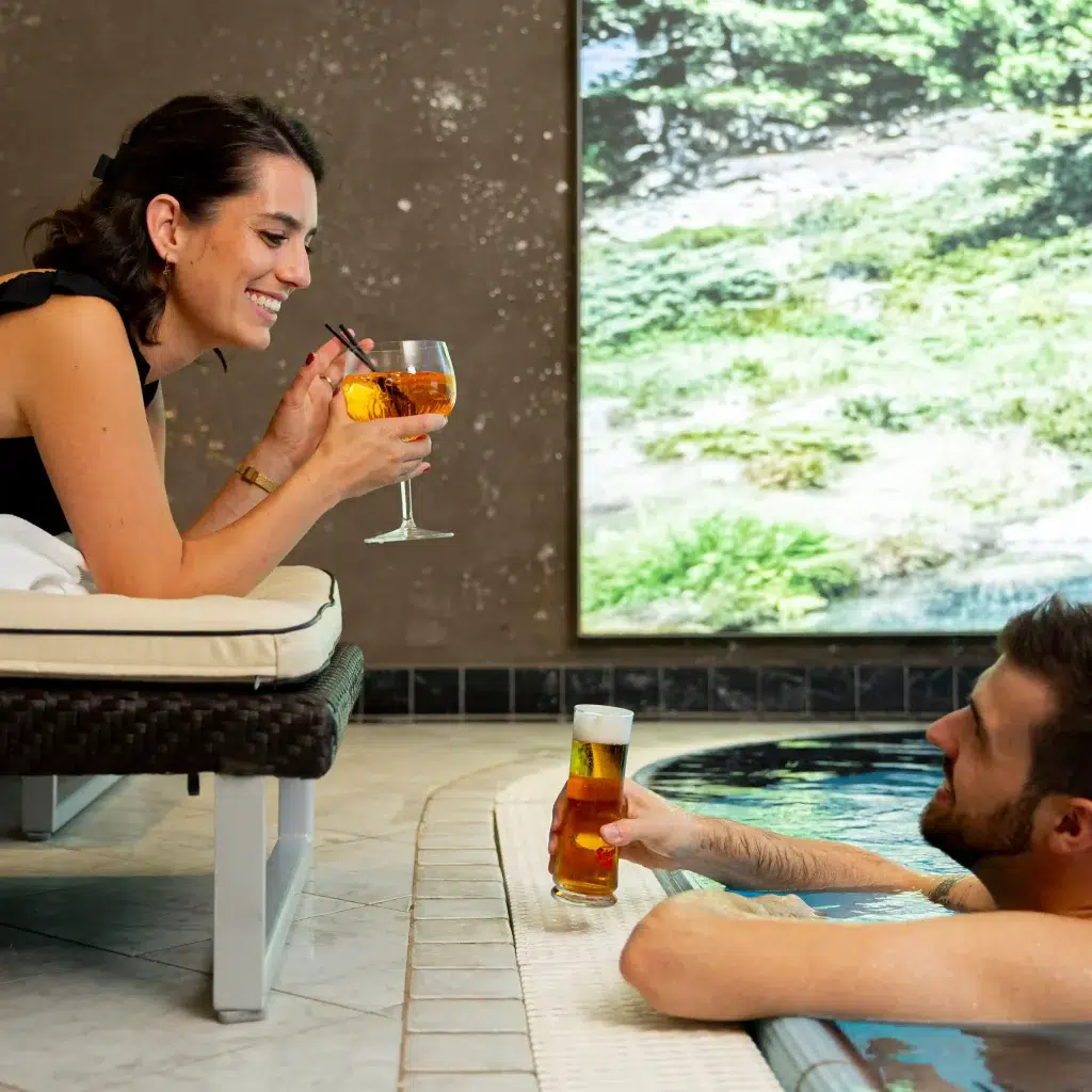 A woman reclines on a lounge chair, smiling and holding a cocktail. A man in the pool smiles back, holding a beer. They toast together. A large nature photograph decorates the wall behind them.