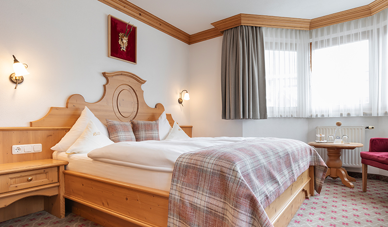 A cozy single room in Ischgl featuring a wooden bed with white and plaid bedding. Two wall lamps flank the bed, while a small round table with chairs sits near a window adorned with white and gray curtains. The room boasts a light color scheme and patterned carpet.