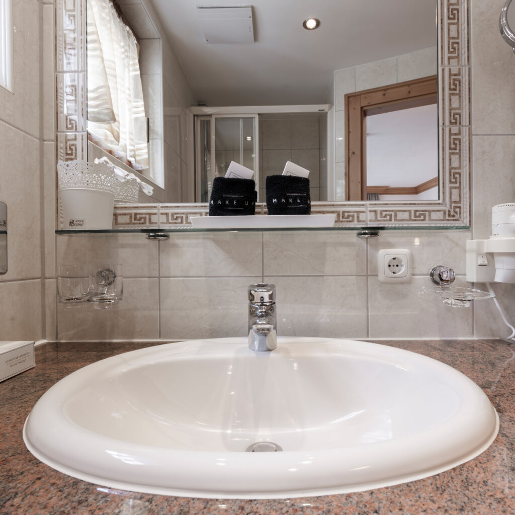 Dieses moderne Einzelzimmer in Ischgl verfügt über ein elegantes Badezimmer mit weißem Waschbecken und bronzefarbener Waschtischplatte. Ein großer Spiegel mit dekorativen Fliesen spiegelt den Duschbereich und die Holzakzente wider, während schwarze Toilettenartikelhalter und zwei gefaltete Handtücher auf einem Glasregal darüber ruhen.