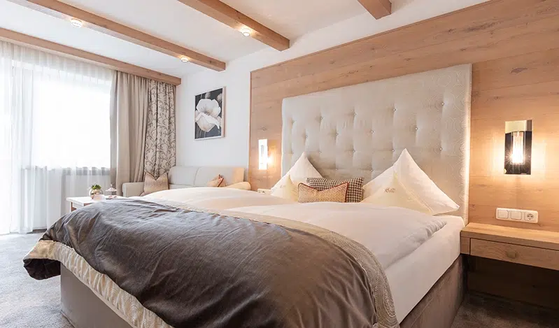 A cozy Suite Ischgl bedroom with a large bed featuring a tufted headboard and soft bedding in neutral tones. Wooden beams on the ceiling and a wooden accent wall add warmth. A small sofa and floral artwork are visible near the window with sheer curtains.
