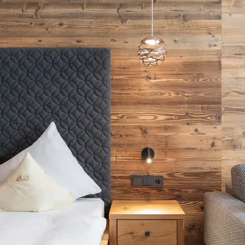Dieses Doppelzimmer Ischgl verfügt über ein gemütliches Schlafzimmer mit einem modernen, strukturierten grauen Kopfteil und einer Holzwand. Ein kleiner Nachttisch aus Holz mit einer Schublade hält eine Lampe, während darüber eine dekorative Pendelleuchte hängt. Weiße Kissen und Laken verleihen einen eleganten Hauch von Schlichtheit.