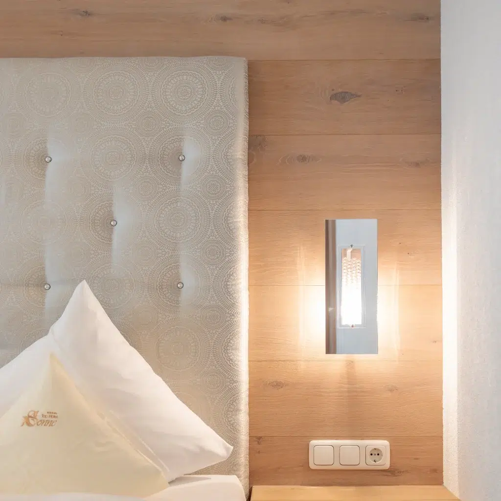 Nahaufnahme eines modernen Suite Ischgl-Schlafzimmers mit gepolstertem Kopfteil, einem ordentlich arrangierten Kissen mit aufgesticktem Logo und einer eleganten Wandleuchte. Die Wand ist mit hellem Holz verkleidet und hat zwei Steckdosen unter der Leuchte.