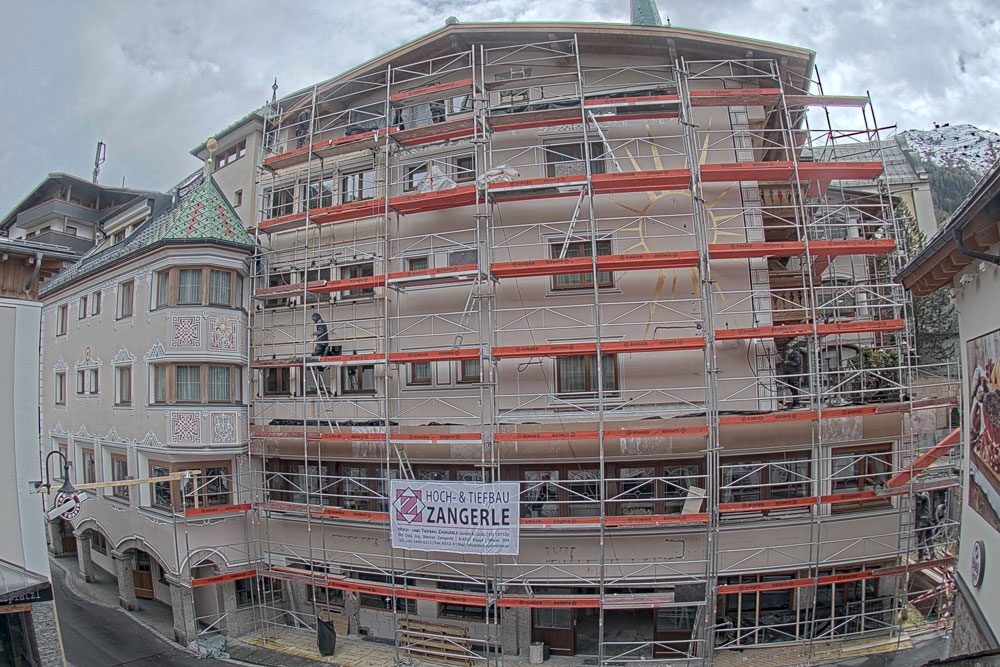 A large building is covered in scaffolding with a banner that reads HOCH- & TIEFBAU ZANGERLE. A construction worker is visible on the scaffold. The surrounding street and adjacent buildings have an alpine architectural style.