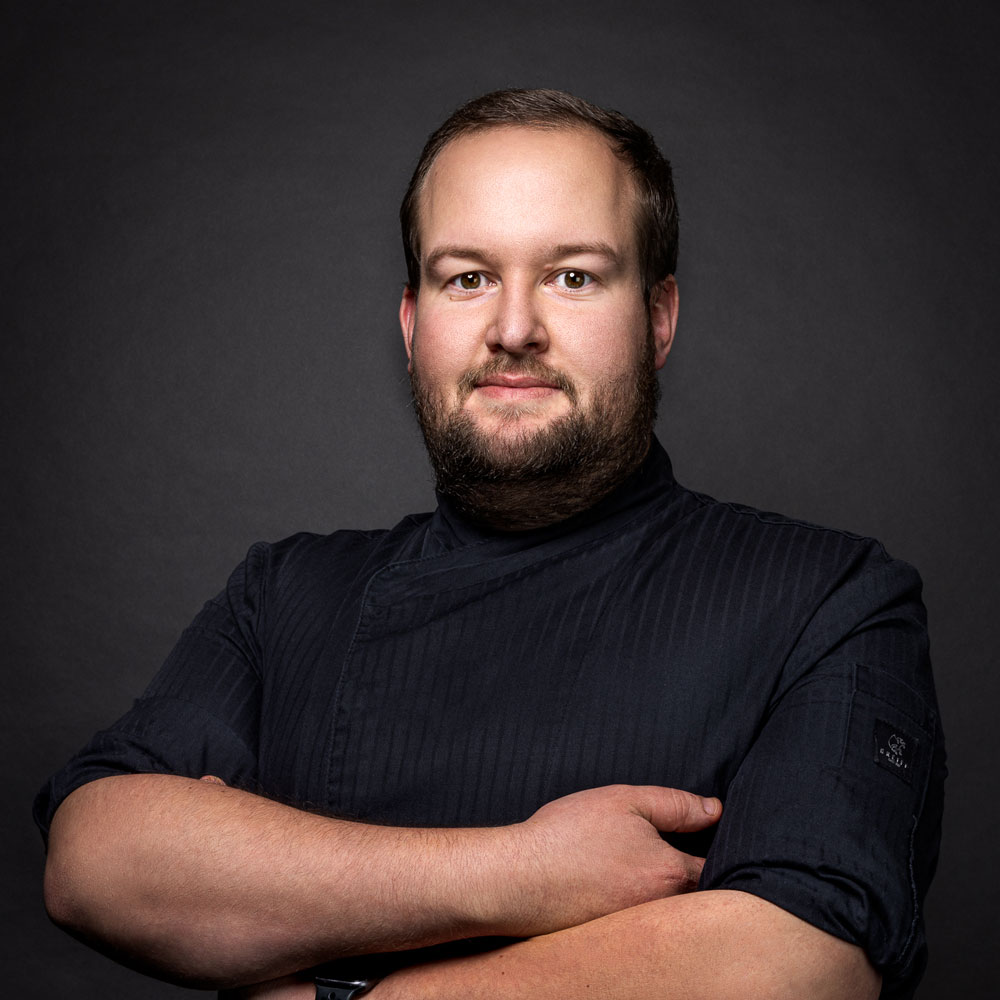 A man with a beard, wearing a dark chefs jacket, stands with arms crossed against a dark background.