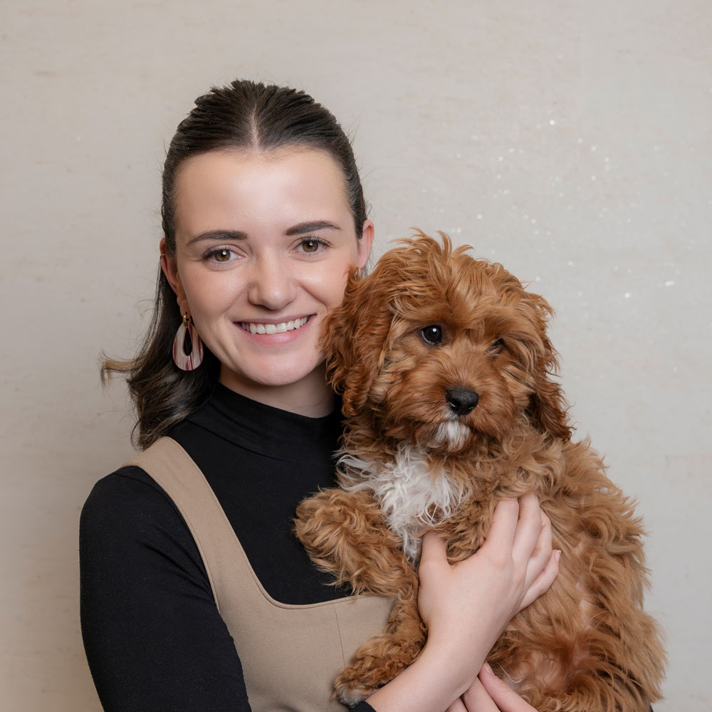 A smiling person with dark hair, wearing a black top and beige jumper, holds a small, curly-haired brown dog. The background is plain and light-colored.