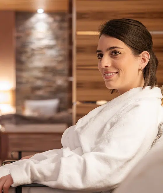 Eine Frau mit dunklem Haar sitzt lächelnd in einer gemütlichen Hotelsuite in Ischgl. Sie trägt einen weißen Bademantel und strahlt Wohlbefinden und Gelassenheit aus. Der Hintergrund ist durch sanftes Licht, eine Steinmauer und Holzdekor geprägt und schafft eine warme und entspannende Atmosphäre.