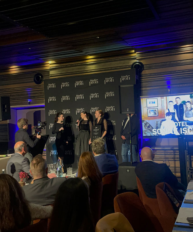 People on stage at an event, some holding microphones. A cameraman films them. The backdrop displays Gastro PROFI. A screen shows a group photo with the word HOTEL SAN FRANCISCO. Audience members watch from seated tables.