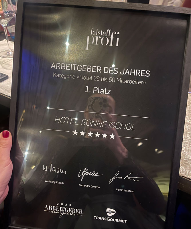 A close-up of a framed award plaque held by a hand with red nail polish. The plaque, titled falstaff profi, recognizes Hotel Sonne Ischgl as the Employer of the Year in the category for hotels with 26 to 50 employees, achieving first place.