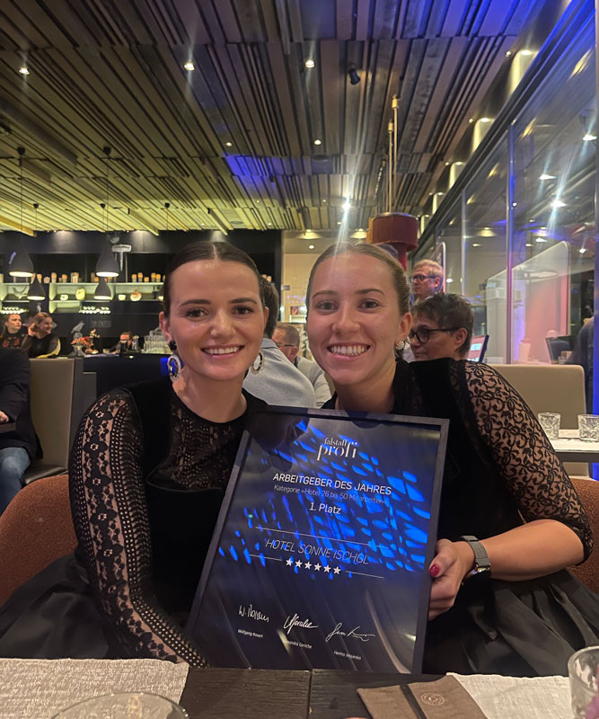 Two smiling women in black outfits sit in a restaurant, holding a glass award with blue accents. The award reads Arbeitgeber des Jahres, 1. Platz, Hotel Schweizerhof. The background features modern decor with people dining.