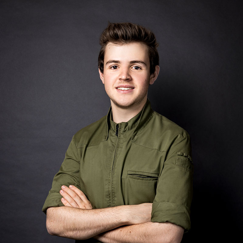A person with short brown hair is smiling at the camera. They are wearing a green zip-up jacket and have their arms crossed. The background is plain and dark, highlighting the individual.