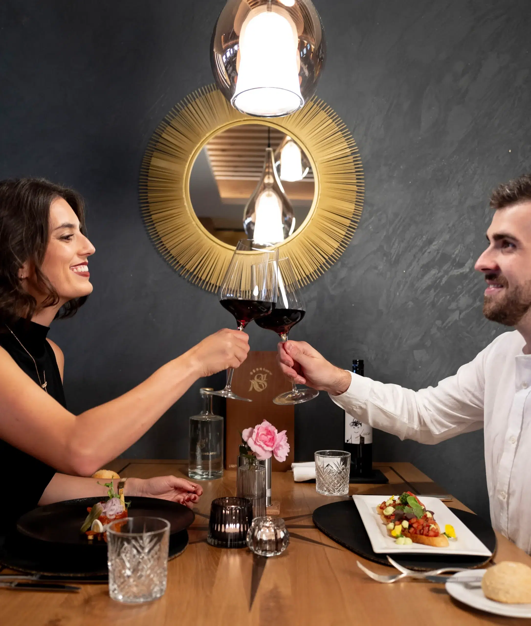 A woman and a man, both smiling, clink glasses of red wine at a gourmethotel in Ischgl. The table is set with elegant dishes, and a decorative golden mirror on the wall behind them reflects the warm lighting.