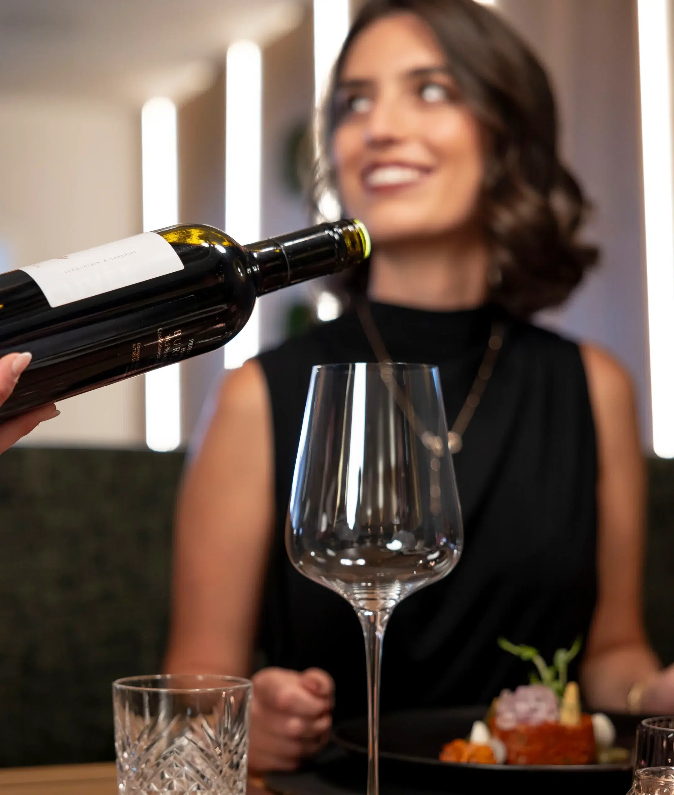 At a gourmet hotel in Ischgl, a person in a black dress sits at a table, smiling, as another hand pours wine into their glass. A plate of food and water are in front of them, with the background softly blurred.