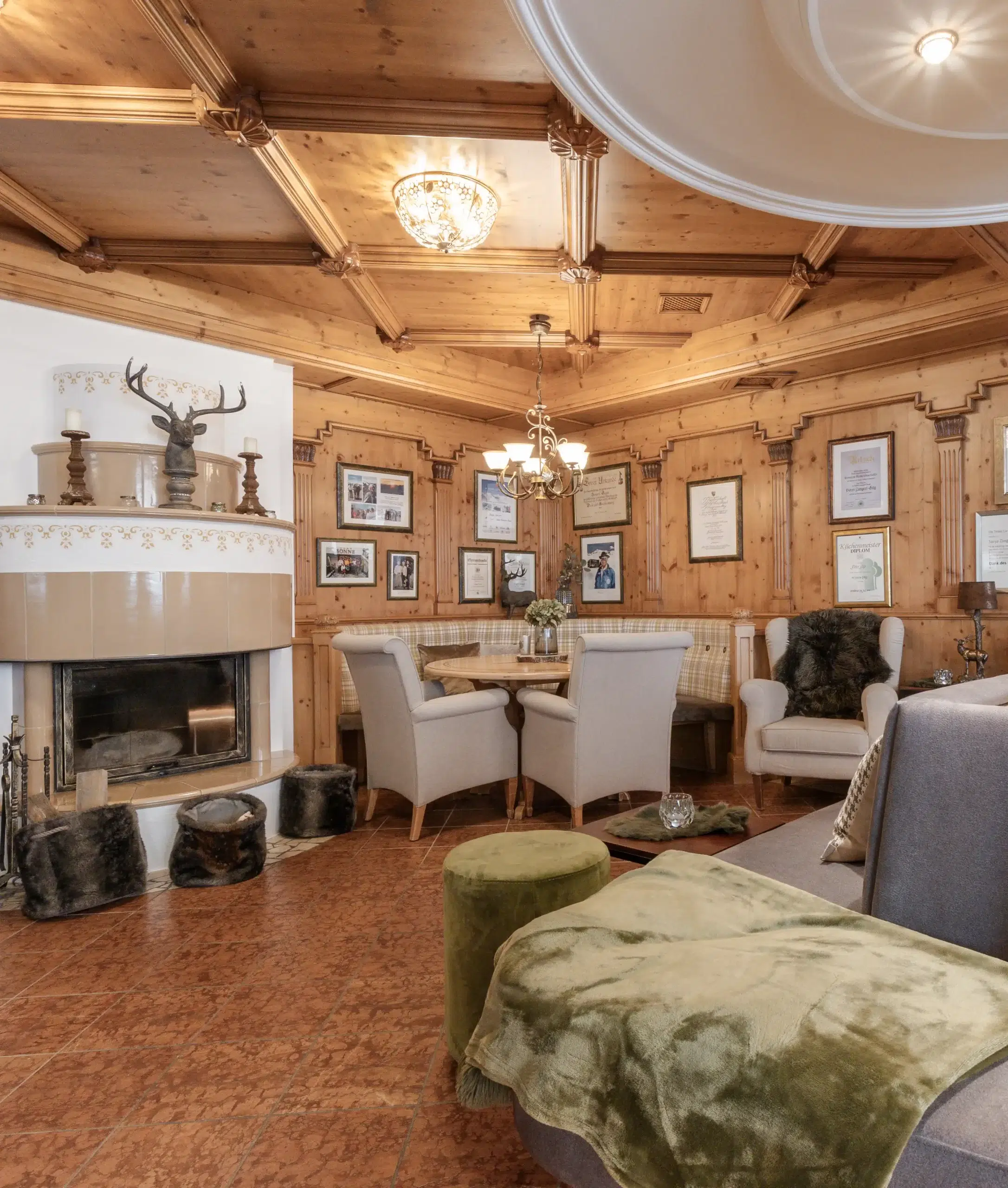 A cozy room with wood-paneled walls and ceiling, reminiscent of Hotelbar Ischgl, featuring a fireplace with a deer head surrounded by framed pictures. Cushioned chairs around a small table, an olive green ottoman, and a soft blanket on the sofa complete this inviting space.