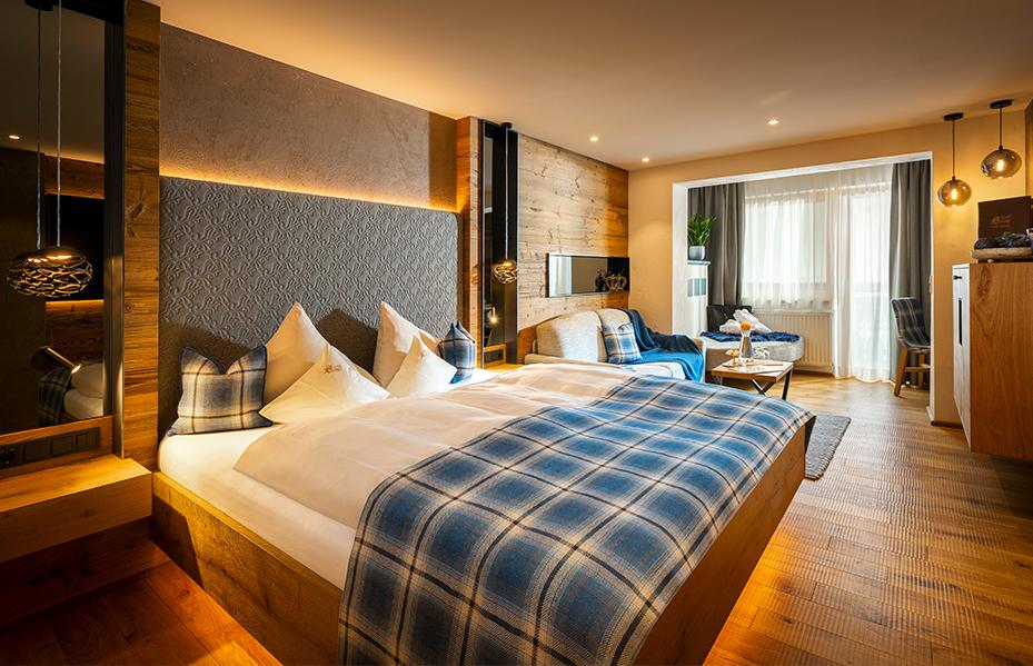 A cozy, modern hotel room with a large bed adorned with plaid bedding and pillows. The room features wooden decor, a wall-mounted TV, a small sitting area by the window, and warm lighting from hanging lamps.