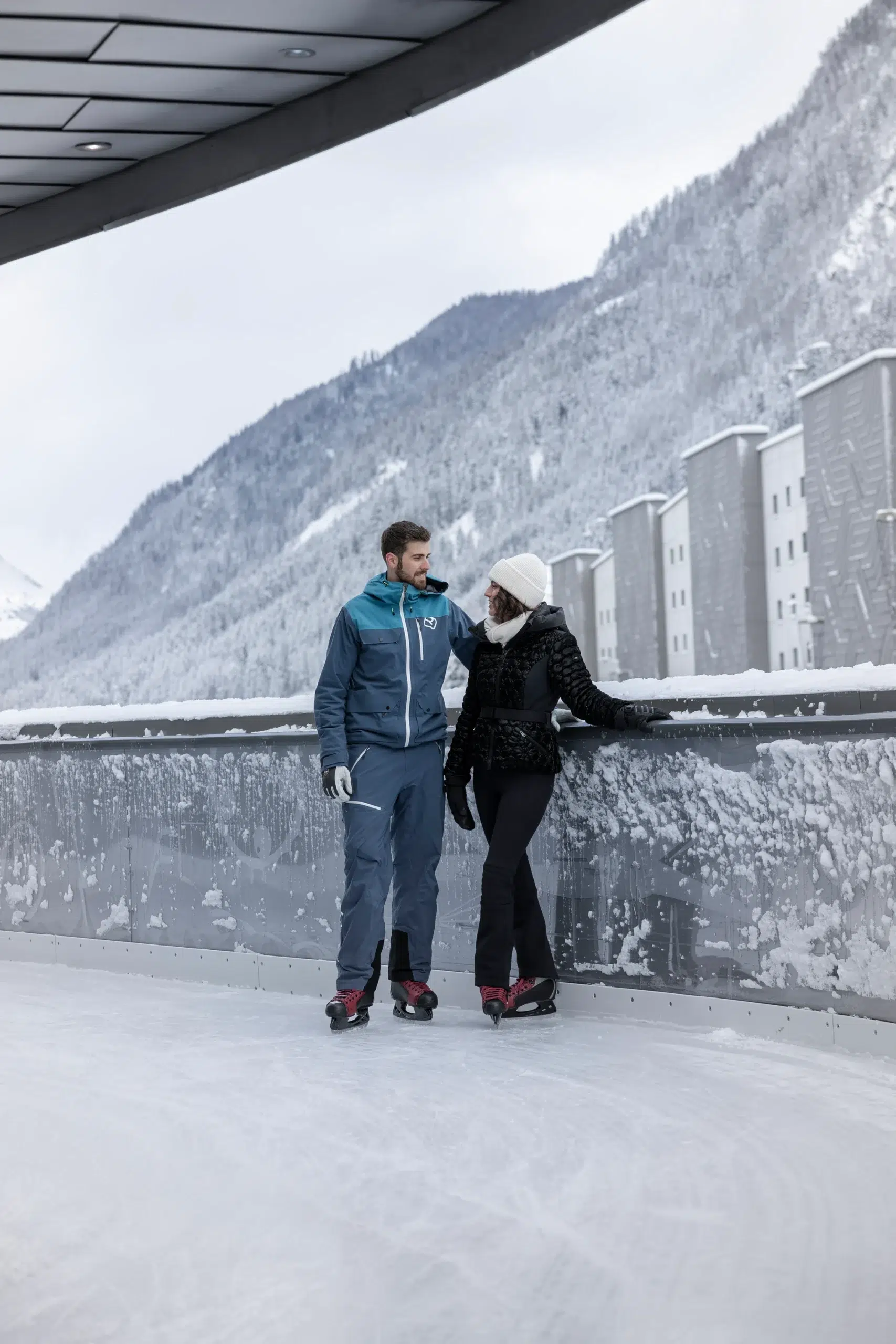 A man and woman, dressed for their winter holiday in Ischgl, stand on an outdoor ice rink in their ice skates. Leaning on a snow-covered railing, snowy mountains and charming buildings frame the scene. The man sports a blue jacket while the woman dons a black jacket and white hat.