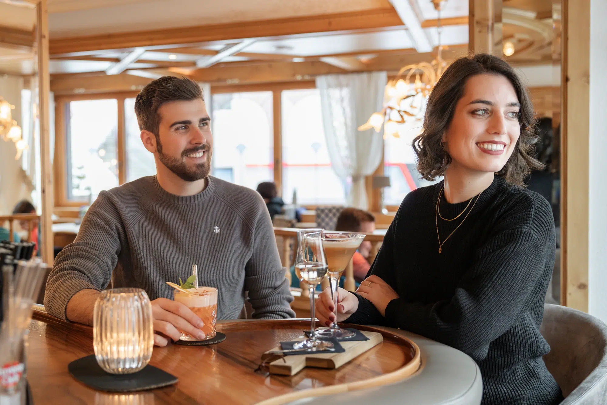 A man and woman sitting at a wooden bar, each holding a drink. The man has a cocktail with a garnish, while the woman holds a glass of wine. They are smiling and looking to their left. The bar is warmly lit and features wooden decor.