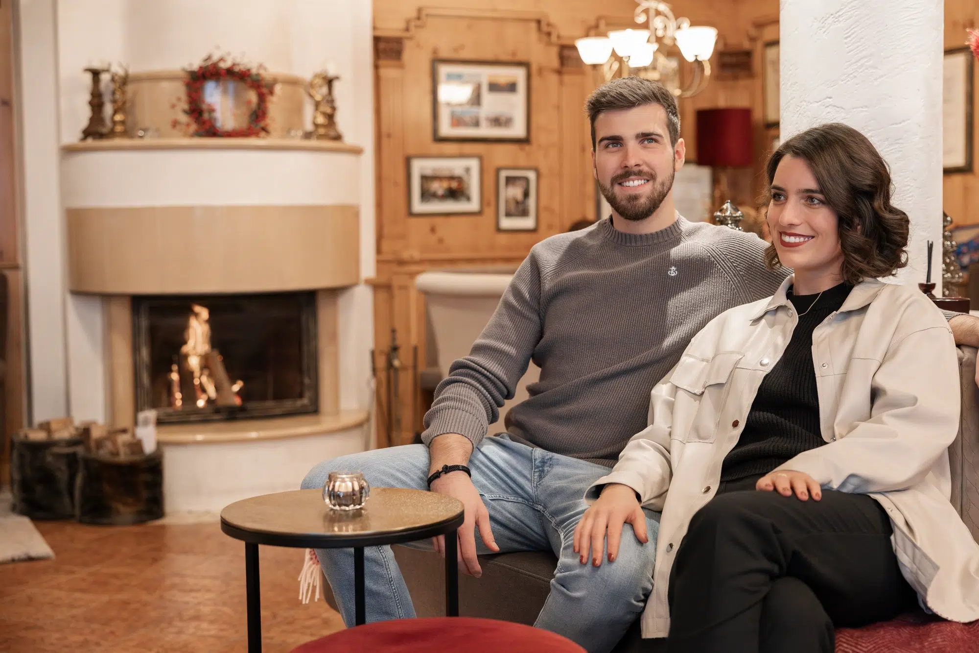 A man and woman sit on a couch in a cozy room with wooden walls and a lit fireplace. They are smiling, and a small, round table with a candle is in front of them. The room is decorated with framed pictures and ornaments.