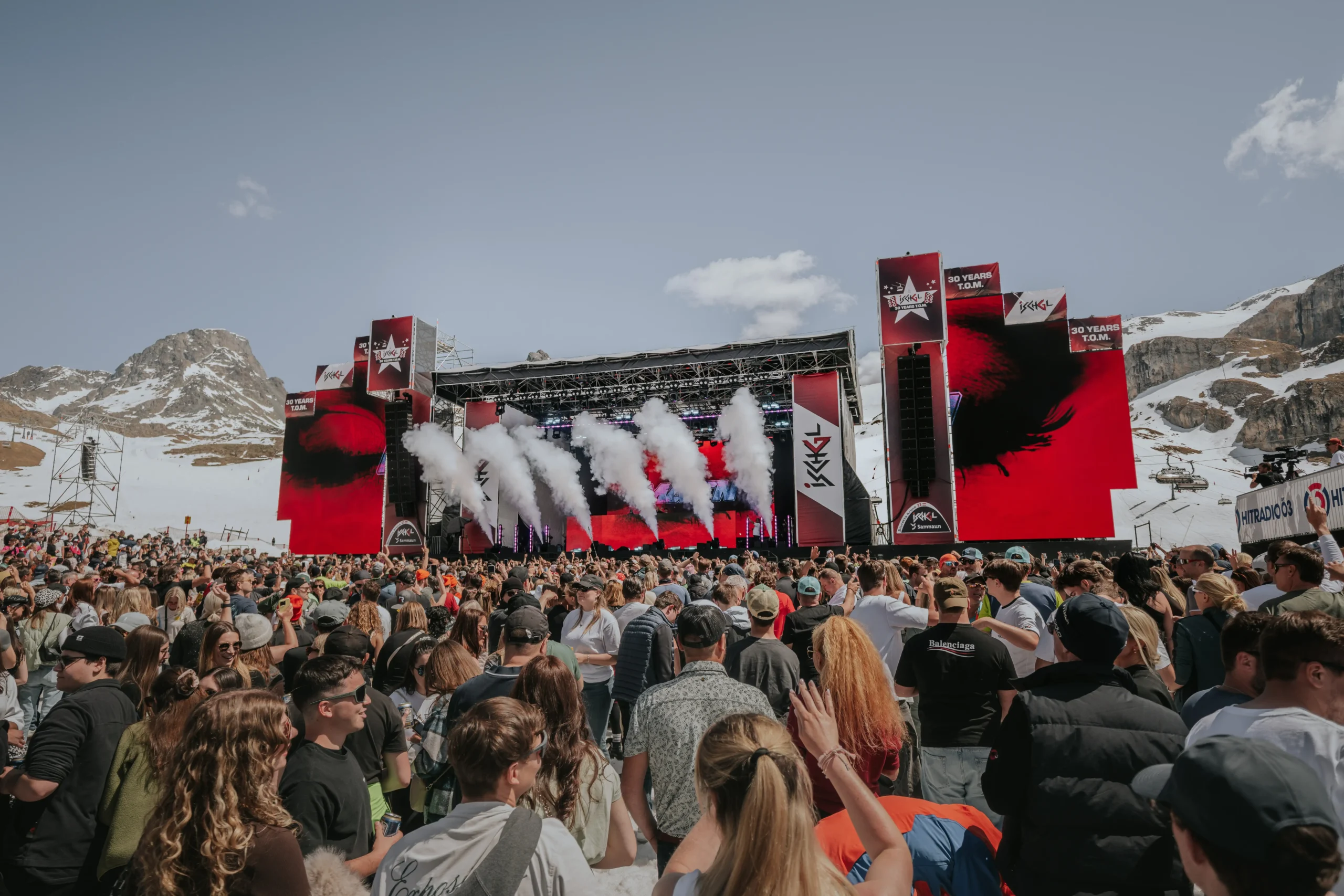 Large crowd at an outdoor concert with mountains in the background and smoke on stage.